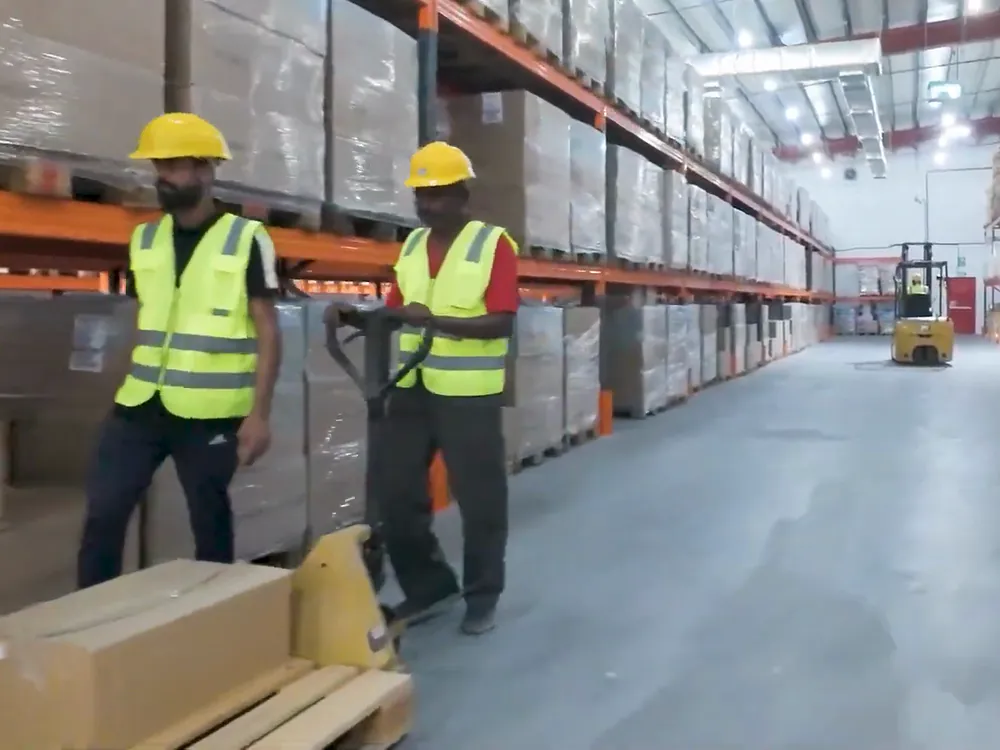 Two Liqa warehouse staff in safety vests and hard hats moving stock with a pallet truck